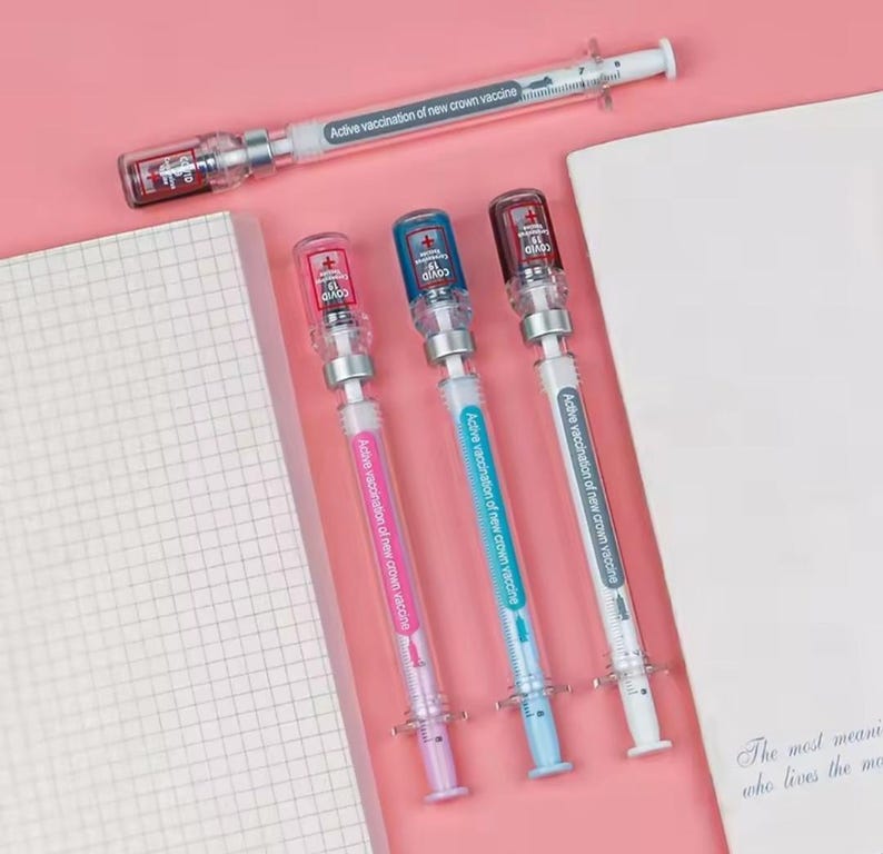 May include: A collection of syringe-shaped pens with colorful accents. The pens have clear barrels with colored plungers and caps, and the text "Active vaccination of new crown vaccine" is printed on the barrels. The pens are arranged on a pink surface.