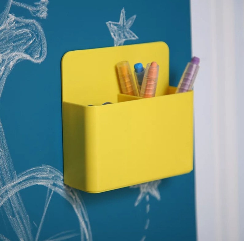 May include: A bright yellow wall-mounted organizer with three compartments, holding colorful markers and pens. The organizer is mounted on a blue surface with white chalk drawings of stars and other shapes.