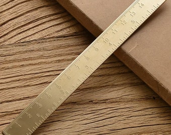 May include: A gold-colored metal ruler with markings from 0 to 15. The ruler is on a wooden surface.