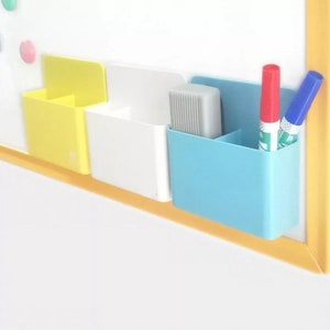 May include: A set of three colorful, magnetic whiteboard organizers. The organizers are yellow, white, and blue, and are attached to a white board. The blue organizer holds a red and blue marker. The white organizer holds a gray eraser.
