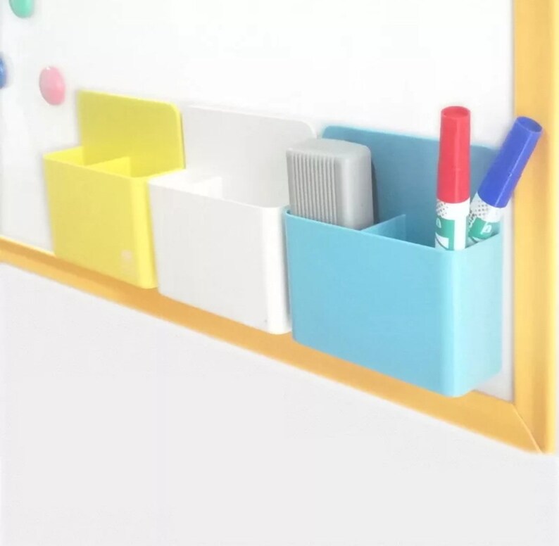 May include: A set of three colorful, magnetic whiteboard organizers. The organizers are yellow, white, and blue, and are attached to a white board. The blue organizer holds a red and blue marker. The white organizer holds a gray eraser.
