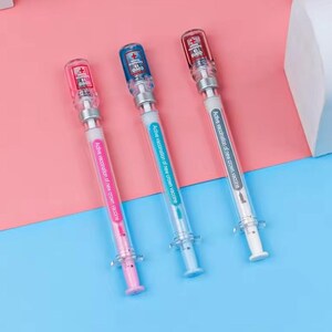 May include: Three syringe-shaped pens with clear barrels and colorful accents. Each pen has a different colored plunger: pink, blue, and red. The pens have the text "COVID 19" and "Active vaccination of new crown vaccine."