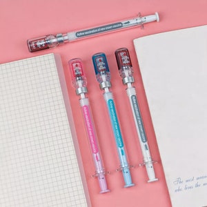 May include: A collection of syringe-shaped pens in pink, blue, and white, with the text "Active vaccination of new crown vaccine." Each pen has a clear barrel and a cap with a COVID-19 logo. One pen is positioned at the top, and three are aligned below.