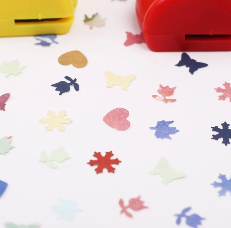 May include: Two red and yellow paper punch tools with a white background. The tools are used to create confetti in the shapes of hearts, butterflies, snowflakes, and leaves. The confetti is scattered around the tools.