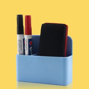 May include: A light blue desk organizer with a black marker, a red marker, and a black eraser. The organizer has three compartments. The markers have white labels with black text. The eraser has a red frame.