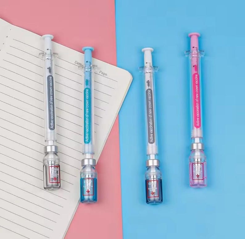 May include: Four syringe-shaped pens with clear barrels and colorful plungers: white, blue, white, and pink. Each pen has a small bottle-shaped base with a cross symbol and the text "Active vaccination of new crown vaccine". The pens are on a blue and pink background.