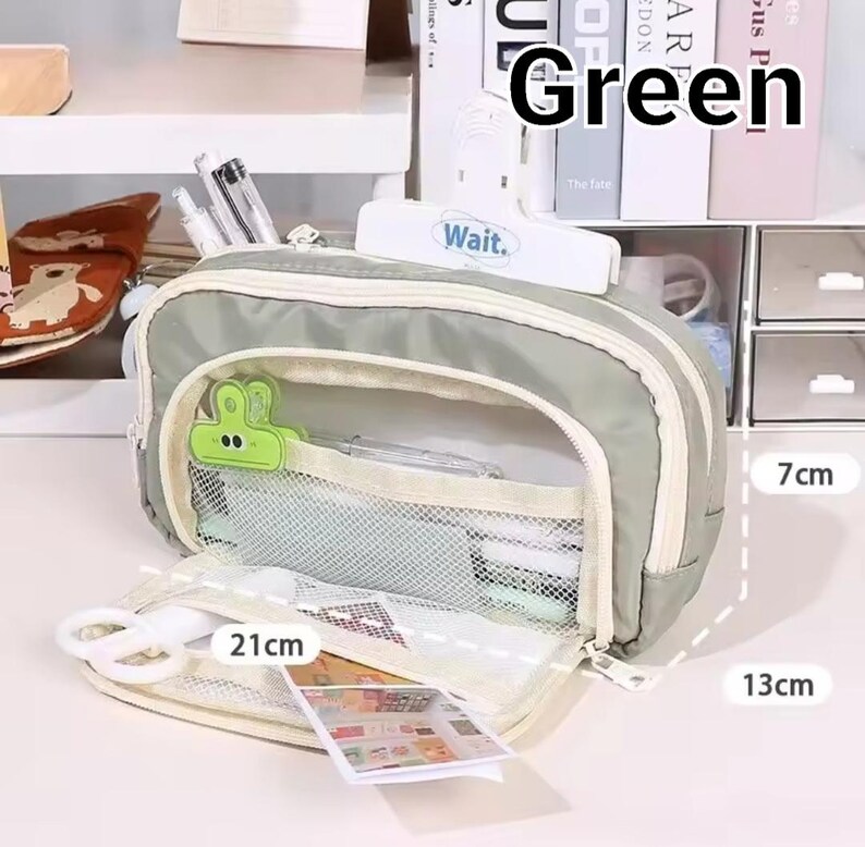 May include: A sage green pencil case with a zippered opening, a white mesh interior pocket, and a green frog-shaped clip. The case measures 21 cm in length, 13 cm in height, and 7 cm in width. It contains pens, scissors, and a small card.