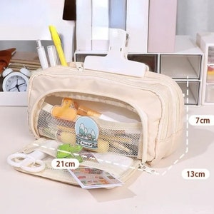 May include: A beige multi-compartment pencil case with a zipper closure, featuring a front mesh pocket containing stationery items. The case measures 21 cm in length, 13 cm in width, and 7 cm in height, with a white clip attached to the top exterior.