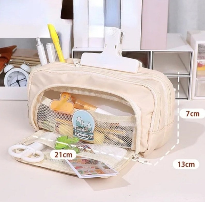May include: A beige multi-compartment pencil case with a zipper closure, featuring a front mesh pocket containing stationery items. The case measures 21 cm in length, 13 cm in width, and 7 cm in height, with a white clip attached to the top exterior.