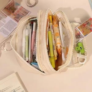 May include: A cream-colored multi-compartment pencil case filled with various stationery items including pens, highlighters, and markers. The open case reveals organized sections containing writing instruments in shades of yellow, white, and pink, alongside a small green clip.