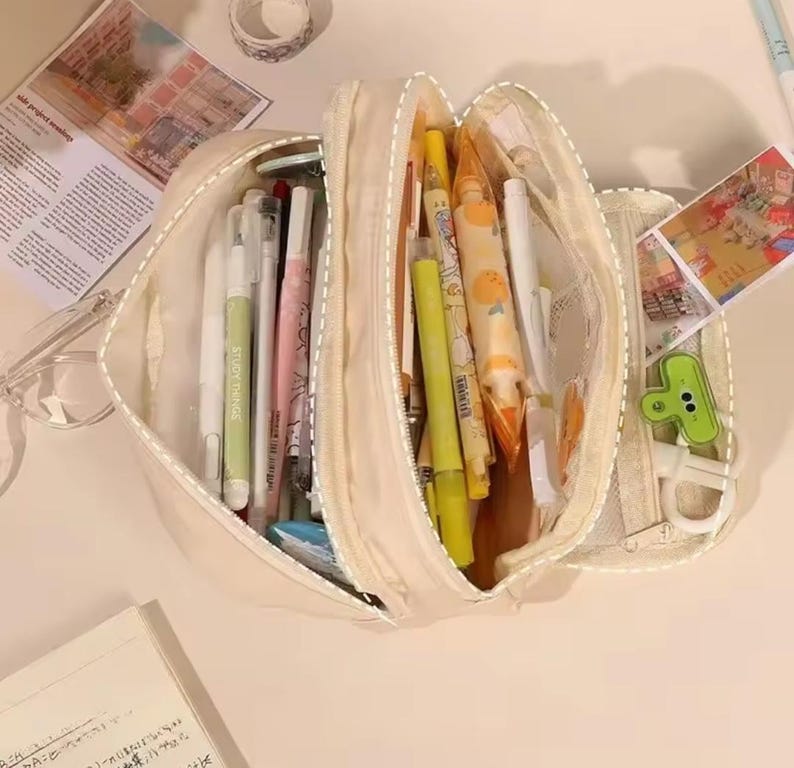 May include: A cream-colored multi-compartment pencil case filled with various stationery items including pens, highlighters, and markers. The open case reveals organized sections containing writing instruments in shades of yellow, white, and pink, alongside a small green clip.