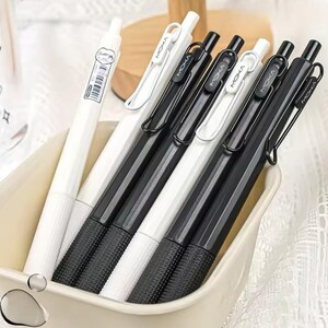 Puede incluir: Una colección de bolígrafos blancos y negros con la palabra "MOKA" impresa en el clip. Los bolígrafos están en un recipiente rectangular de color crema. Los bolígrafos tienen un agarre texturizado y un diseño elegante.