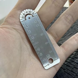 Puede incluir: Una pequeña regla de metal plateado con medidas en pulgadas y centímetros. La regla tiene un extremo curvo con marcas de grados de 0 a 90. La regla mide aproximadamente 12,7 cm de largo.