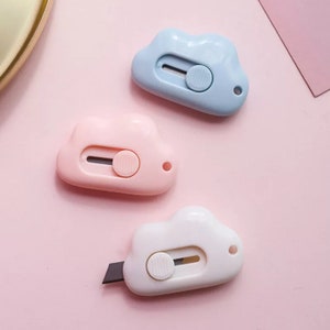 Puede incluir: Tres cuchillos de uso general en forma de nube en rosa, azul y blanco. Cada cuchillo tiene una cuchilla retráctil y un pequeño agujero para colgar.