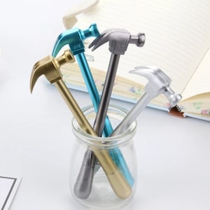 May include: A selection of hammer-shaped pens in a clear glass jar. The pens are in various metallic colours, including gold, silver, and teal. The pens are resting in a clear glass jar, with a notebook in the background.