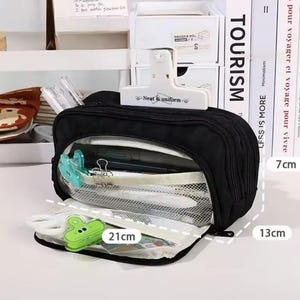 May include: A black multi-compartment pencil case with a white clip labeled Neat & uniform. The case features a mesh interior pocket, a green clip, and white scissors. Dimensions shown are 21 cm in length, 13 cm in height, and 7 cm in depth.