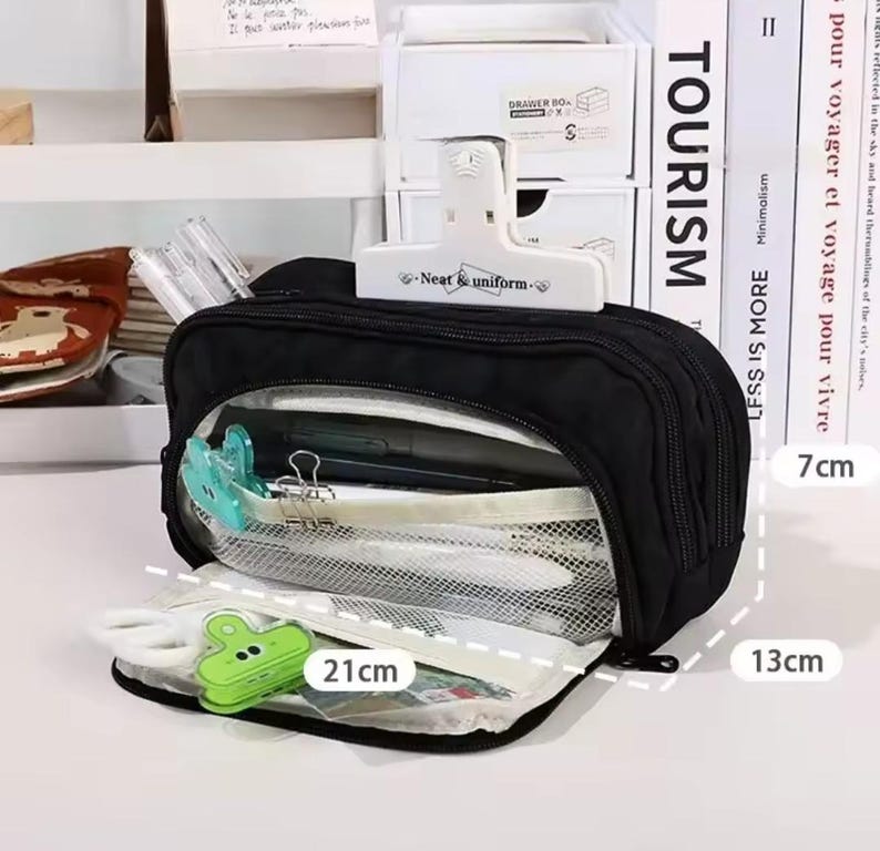 May include: A black multi-compartment pencil case with a white clip labeled Neat & uniform. The case features a mesh interior pocket, a green clip, and white scissors. Dimensions shown are 21 cm in length, 13 cm in height, and 7 cm in depth.