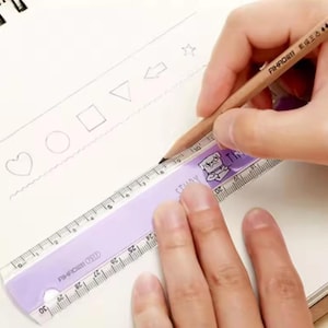 May include: A light purple plastic ruler with a 30 cm scale and a cartoon pug illustration, positioned on a white page with hand-drawn shapes including a heart, circle, square, triangle, arrow, and star, being used with a wooden pencil.