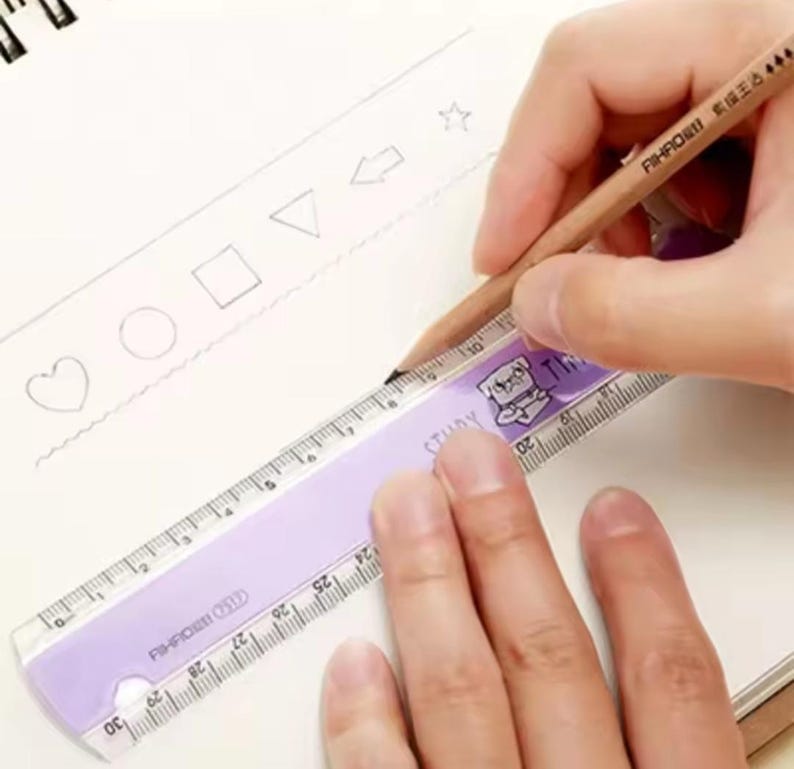 May include: A light purple plastic ruler with a 30 cm scale and a cartoon pug illustration, positioned on a white page with hand-drawn shapes including a heart, circle, square, triangle, arrow, and star, being used with a wooden pencil.