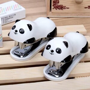 May include: Two white and black panda-shaped staplers with chrome staples. The staplers have a cute panda face design and sit on a light-colored wooden surface. A box with text is visible in the background.