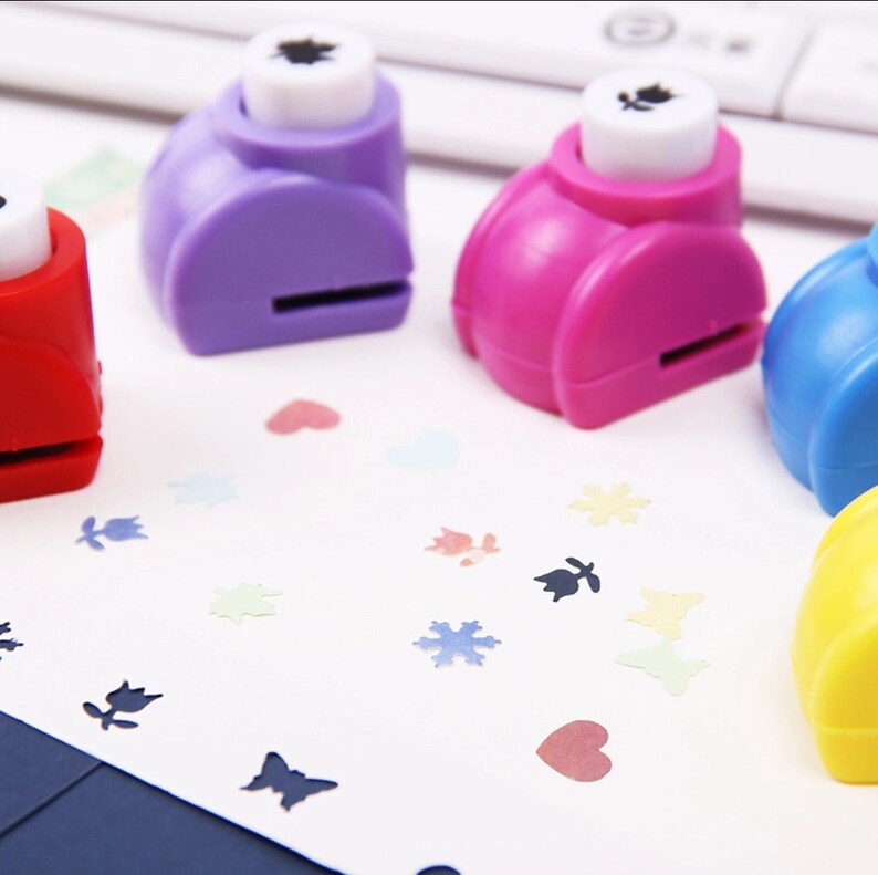 May include: Five colorful paper punch tools with different shapes, including a heart, a snowflake, a leaf, a butterfly, and a flower. The punches are on a white surface with paper cutouts of the shapes.