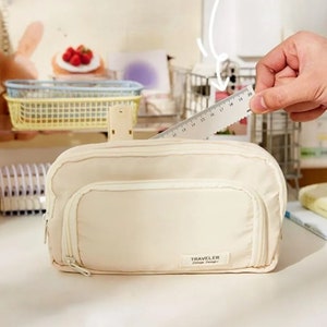 May include: A cream-colored rectangular pencil case with a front zippered pocket and a small rectangular label reading TRAVELER Vintage Design. A clear plastic ruler is positioned behind the case, showing a length of approximately 8 inches.