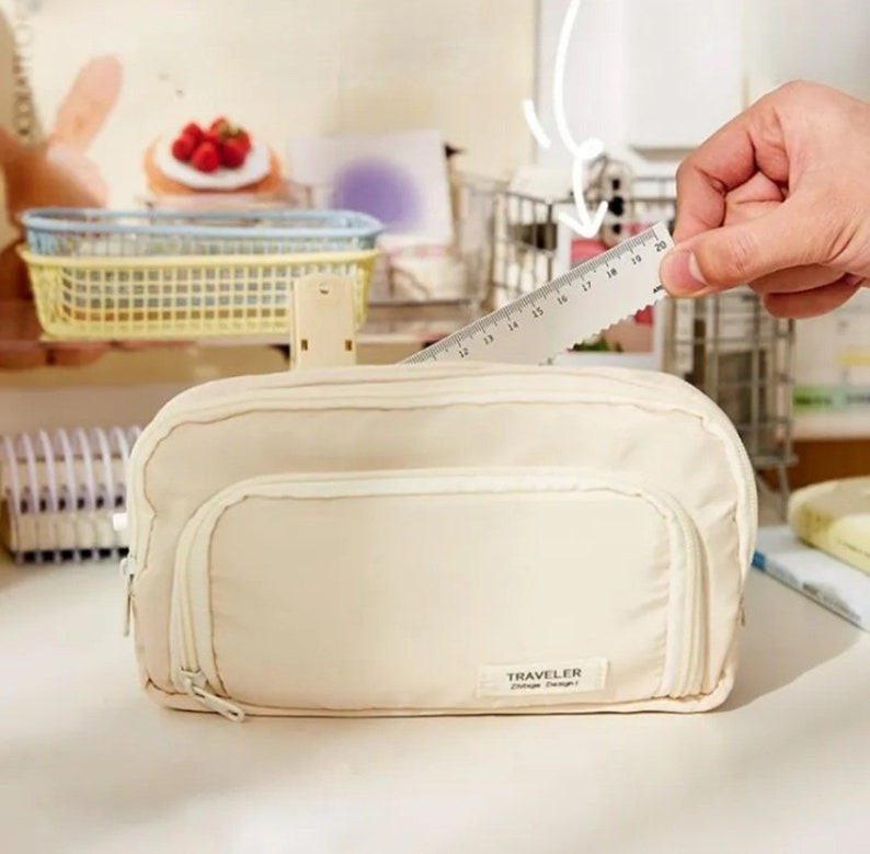 May include: A cream-colored rectangular pencil case with a front zippered pocket and a small rectangular label reading TRAVELER Vintage Design. A clear plastic ruler is positioned behind the case, showing a length of approximately 8 inches.