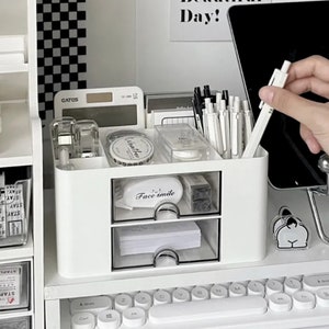 Könnte beinhalten: Weißer zweistöckiger Schreibtisch-Organizer mit durchsichtigen Schubladen. Die obere Schublade enthält einen Hefter, einen Klebebandspender und Stifte. Die untere Schublade enthält einen kleinen Notizblock und andere Büroartikel. Der Organizer steht auf einem weißen Schreibtisch mit einer Tastatur im Vordergrund.