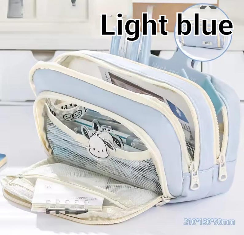 May include: A light blue multi-compartment pencil case with white zippers and trim. The front features a mesh pocket with a cartoon character graphic and a small notebook. The dimensions are printed as 210*150*90mm. The text Light blue is visible.