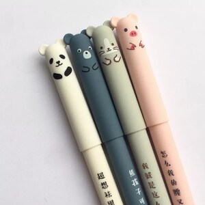 Peut inclure: Quatre stylos colorés avec des embouts en forme d'animaux. Les stylos sont blancs, bleus, gris et roses. Les embouts en forme d'animaux sont un panda, un ours, un chat et un cochon. Les stylos ont des caractères chinois écrits dessus.