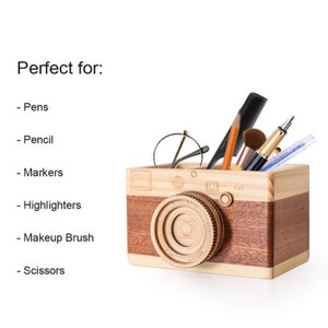 May include: Wooden camera-shaped desk organizer with a brown and natural wood finish. The organizer is filled with various items, including pens, pencils, a comb, makeup brushes, and glasses.