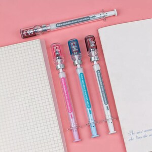 May include: A collection of syringe-shaped pens in pink, blue, and white, with the text "Active vaccination of new crown vaccine." Each pen has a clear barrel and a cap with a COVID-19 logo. One pen is positioned at the top, and three are aligned below.