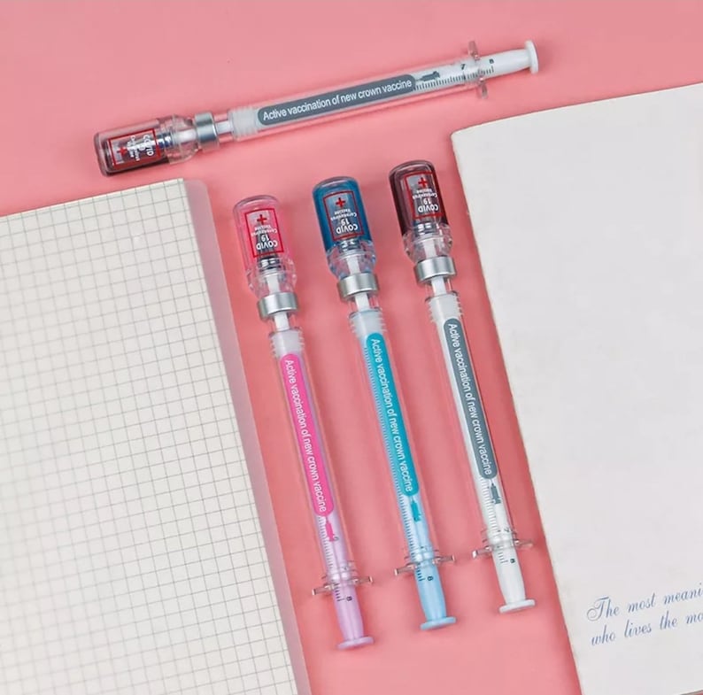 May include: A collection of syringe-shaped pens in pink, blue, and white, with the text "Active vaccination of new crown vaccine." Each pen has a clear barrel and a cap with a COVID-19 logo. One pen is positioned at the top, and three are aligned below.