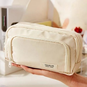 May include: A rectangular, cream-colored zippered pouch featuring a front pocket and a small rectangular fabric label on the lower right corner with the text TRAVELER ZNtage Design. The pouch has a main zipper closure and a secondary front zipper.