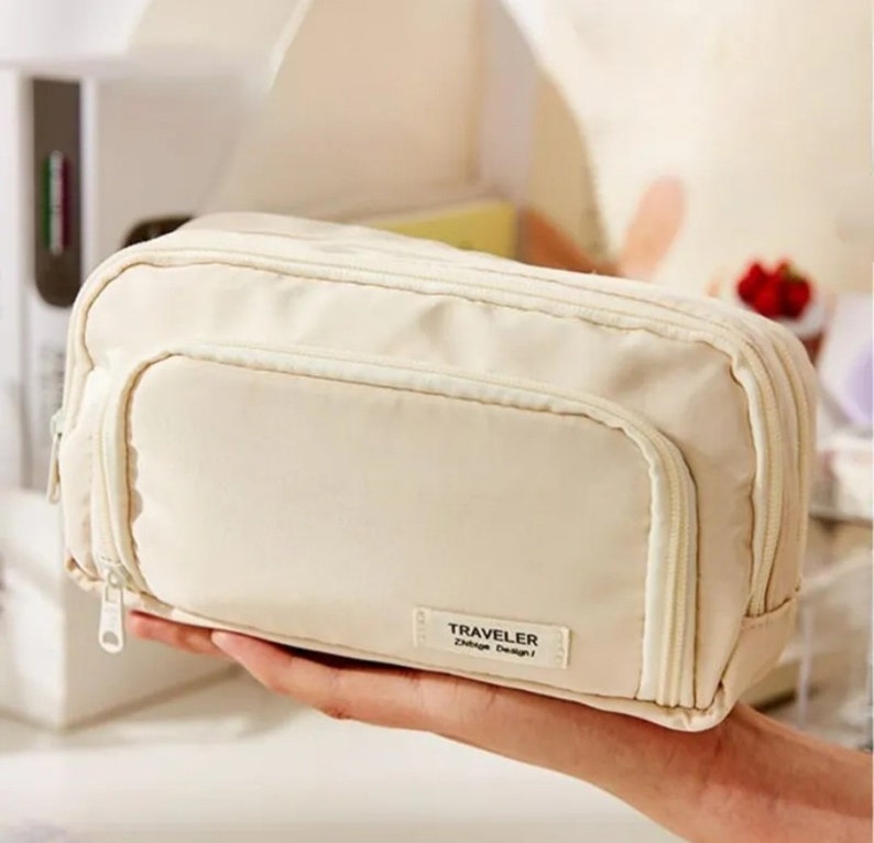 May include: A rectangular, cream-colored zippered pouch featuring a front pocket and a small rectangular fabric label on the lower right corner with the text TRAVELER ZNtage Design. The pouch has a main zipper closure and a secondary front zipper.