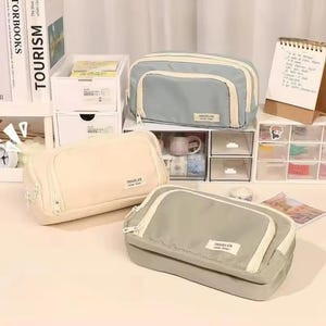 May include: Three rectangular zippered pouches in light blue, cream, and sage green. Each pouch features a front pocket with a white zipper and a small rectangular label on the front that reads TRAVELER Vintage Design in black text.