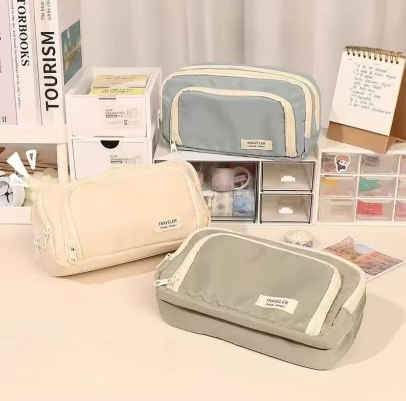 May include: Three rectangular zippered pouches in light blue, cream, and sage green. Each pouch features a front pocket with a white zipper and a small rectangular label on the front that reads TRAVELER Vintage Design in black text.