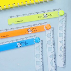 May include: Three plastic folding rulers in lime green, orange, and blue, each featuring a cartoon cat illustration and the text "STUDY TIME!". The rulers have a flower-shaped hinge and include clear sections with various geometric shape cutouts.