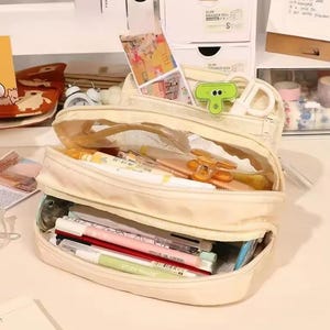 May include: A cream-colored multi-compartment pencil case with a zipper closure, containing various pens, markers, and a green clip. The case features a transparent mesh section and is filled with assorted stationery items, including a light green pen.