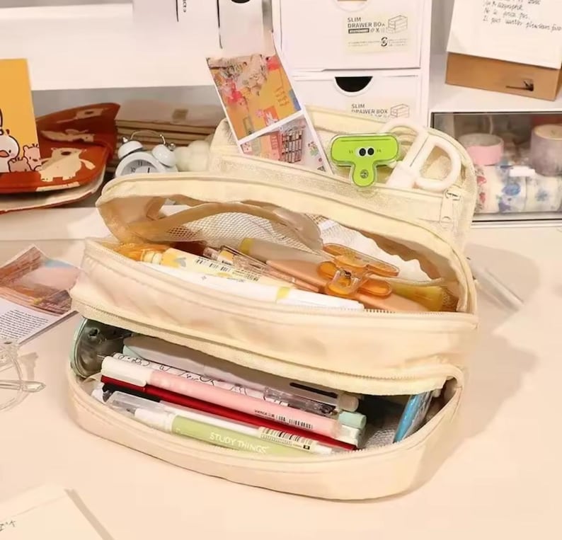 May include: A cream-colored multi-compartment pencil case with a zipper closure, containing various pens, markers, and a green clip. The case features a transparent mesh section and is filled with assorted stationery items, including a light green pen.