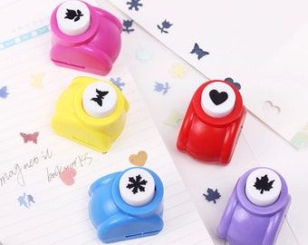 May include: Five colorful paper punchers with different shapes: a flower, a butterfly, a heart, a snowflake, and a leaf. The punchers are on a white and gray notebook with confetti scattered around.