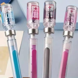 May include: Four clear pens with syringe-like designs. The pens have silver-colored metal accents and are filled with blue, pink, and gray ink. The tops of the pens have a red cross symbol and the words "COVID-19 Vaccine".