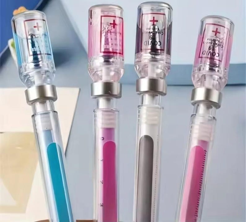 May include: Four clear pens with syringe-like designs. The pens have silver-colored metal accents and are filled with blue, pink, and gray ink. The tops of the pens have a red cross symbol and the words "COVID-19 Vaccine".