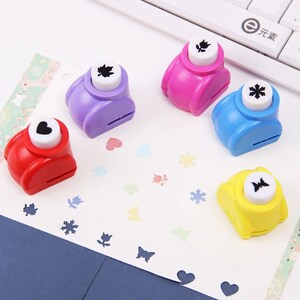 May include: Five colorful paper punchers with different shapes: a heart, a leaf, a flower, a snowflake, and a butterfly. Each puncher has a white button with a black shape on it.