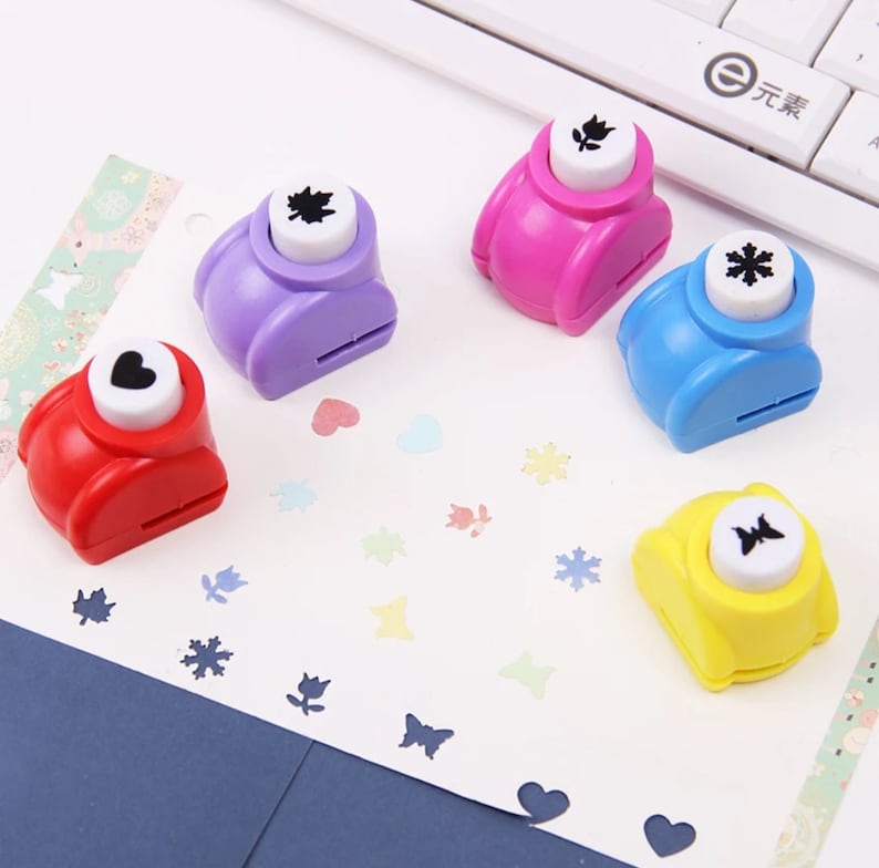 May include: Five colorful paper punchers with different shapes: a heart, a leaf, a flower, a snowflake, and a butterfly. Each puncher has a white button with a black shape on it.