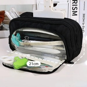 May include: A black rectangular pencil case with multiple zippered compartments and a mesh interior pocket. The case features a white label indicating a length of 21 cm. It is shown open, revealing internal storage for stationery items like pens and clips.