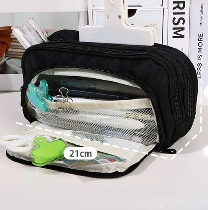 May include: A black rectangular pencil case with multiple zippered compartments and a mesh interior pocket. The case features a white label indicating a length of 21 cm. It is shown open, revealing internal storage for stationery items like pens and clips.