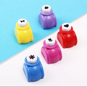 May include: Five colorful paper punchers with different designs: a butterfly, a leaf, a heart, a flower, and a snowflake. The punchers are made of plastic and have a white button with the design on it.
