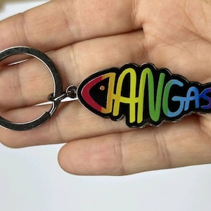 Könnte beinhalten: Ein regenbogenfarbener Fisch-Schlüsselanhänger mit dem Wort "Hangast" darauf. Der Schlüsselanhänger ist aus Metall und hat einen silbernen Ring.