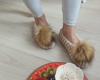 May include: Beige knitted slippers with a large brown fur pom-pom on the top. The slippers are worn by a person wearing light gray leggings. The slippers are on a light wood floor.